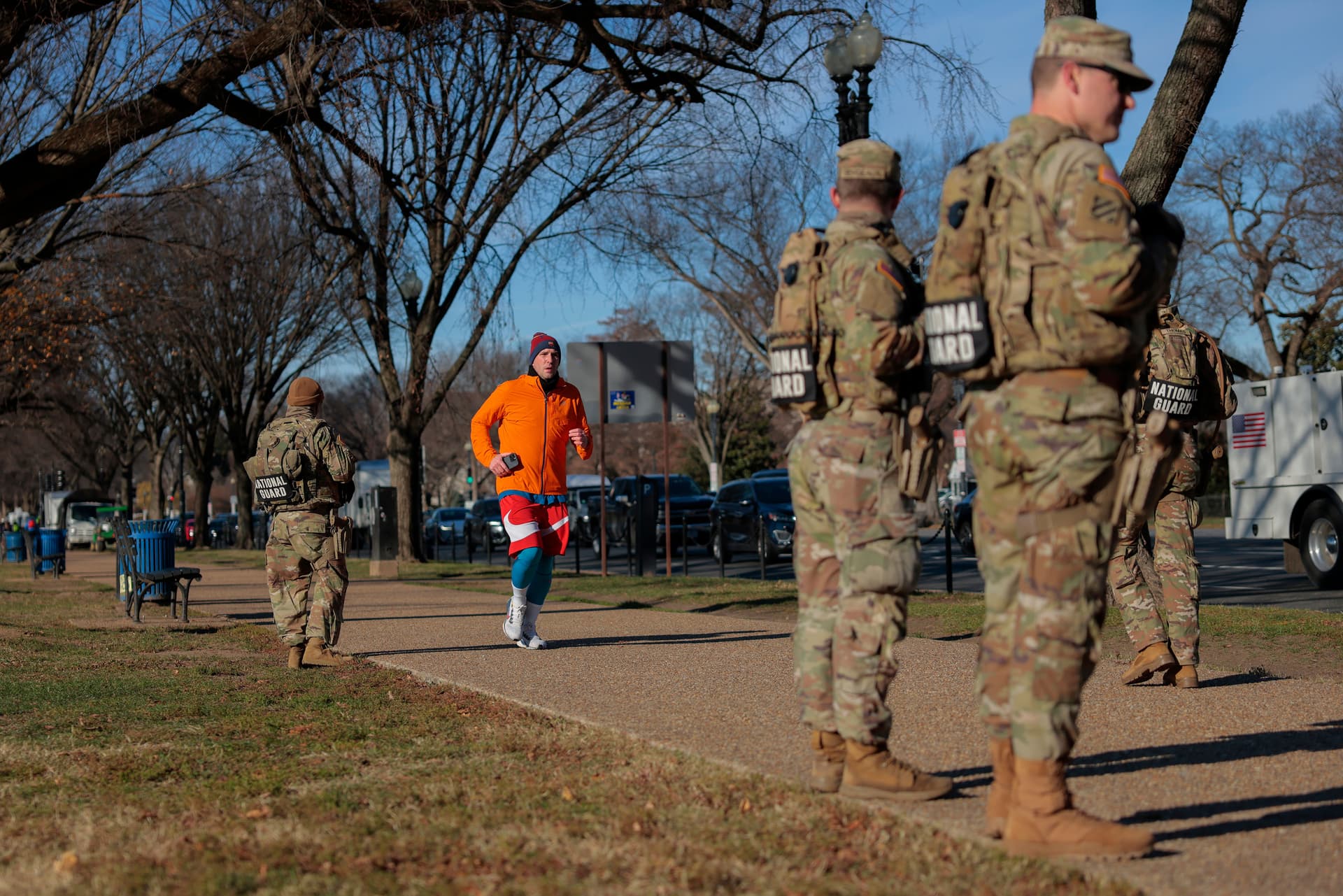 Four members of the National Guard patrols along Constitution Ave. We see their backs as they are standing in rows of two on each side.A runner in an orange and red outfit runs past them.on December 01, 2025 in Washington, DC. Two West Virginia National Guard troops were shot blocks from the White House on November 26, resulting in the death of Sarah Beckstrom on Thursday, November 27, following what authorities called a targeted attack. (