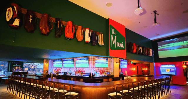 Long L-shaped bar with tvs showing golf. The green wall above the bar displays various golf bags and a sign reading "The Muni"