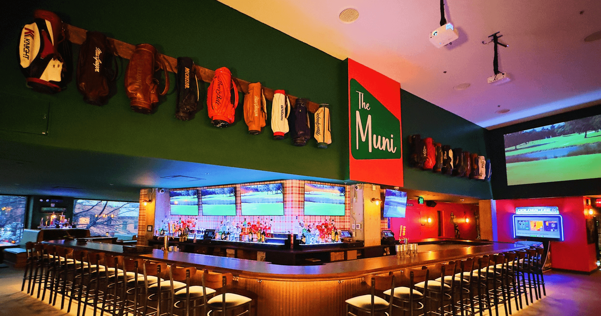 Long, L-shaped bar with sign reading "The muni", several tvs are showing golf and a green wall above the bar displays more than a dozen golf bags